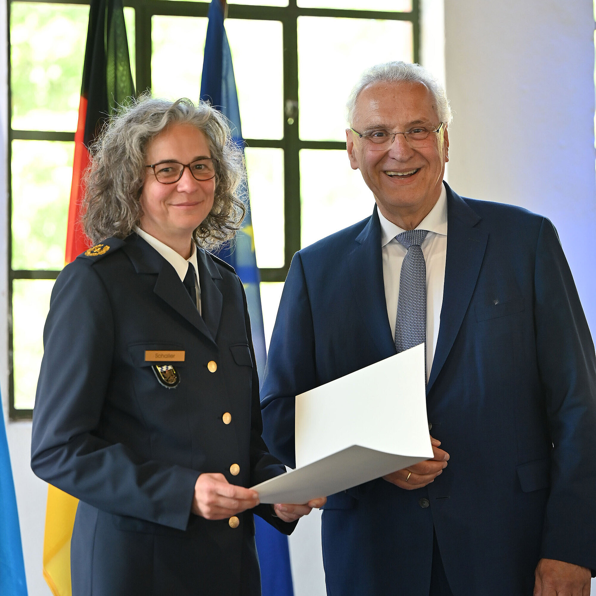 rechts steht  Kerstin Schaller, neue Polizeipräsidentin PP Oberbayern Nord, links Innenminister Joachim Herrmann vor einem Fahnenset, Herrmann überreicht die Ernennungsurkunde