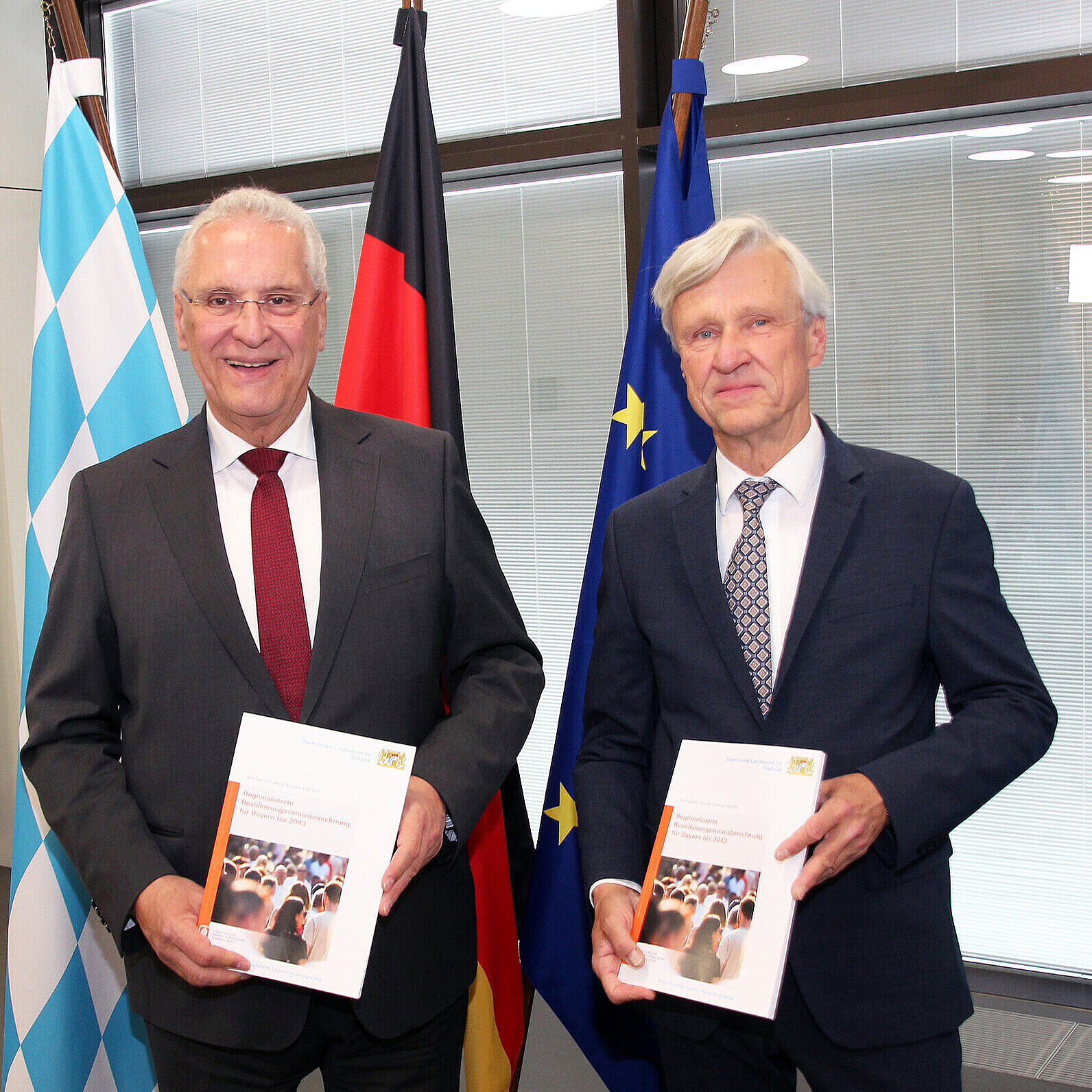 links Innenminister Joachim Herrmann, links Dr. Thomas Gößl, Amtsleiter Bayerisches Landesamt für Statistik stehen vor einem Fahnenset und halten die gebundene Ausgabe Bevölkerungsvorausberechnung 2043 in den Händen
