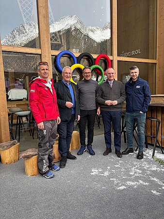 Fünf Personen in Winterkleidung vor einer Holzfassade mit großen bunten Olympischen Ringen; verschneite Berge spiegeln sich im Glas.