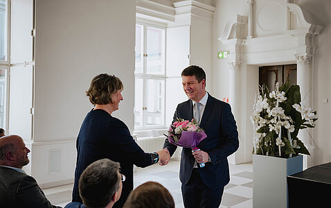 Staatssekretär Sandro Kirchner überreicht einer Frau einen Blumenstrauß und schüttelt ihr die Hand.