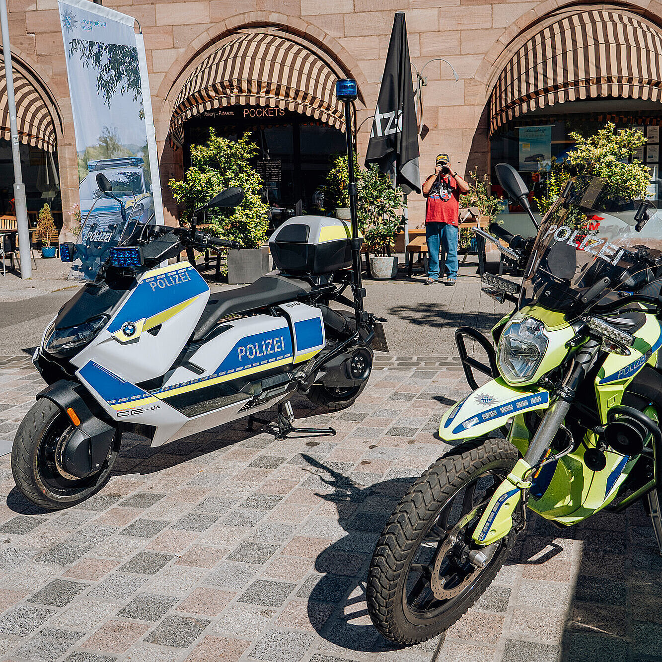 Zwei Polizeifahrzeuge, ein Motorrad und ein Fahrrad, stehen auf einer Stadtstraße. Polizisten in Uniform sind bei der Präsentation der Fahrzeuge anwesend. Ein Mann in einem blauen Anzug steht hinter einem blauen Stand mit der Aufschrift „Polizeipräsidium Mittelfranken“. Im Hintergrund sind Gebäude mit Fenstern und Geschäfte zu sehen.