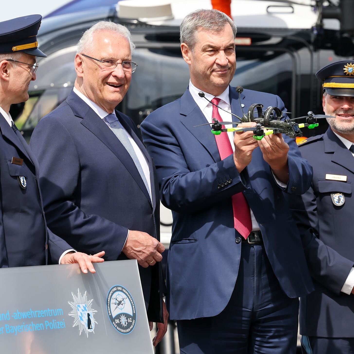 Innenminister Herrmann und Ministerpräsident Söder vor einem Hubschrauber, neben ihnen zwei Polizisten in Dienstuniform. Söder hält eine Mini-Drohne in der Hand.