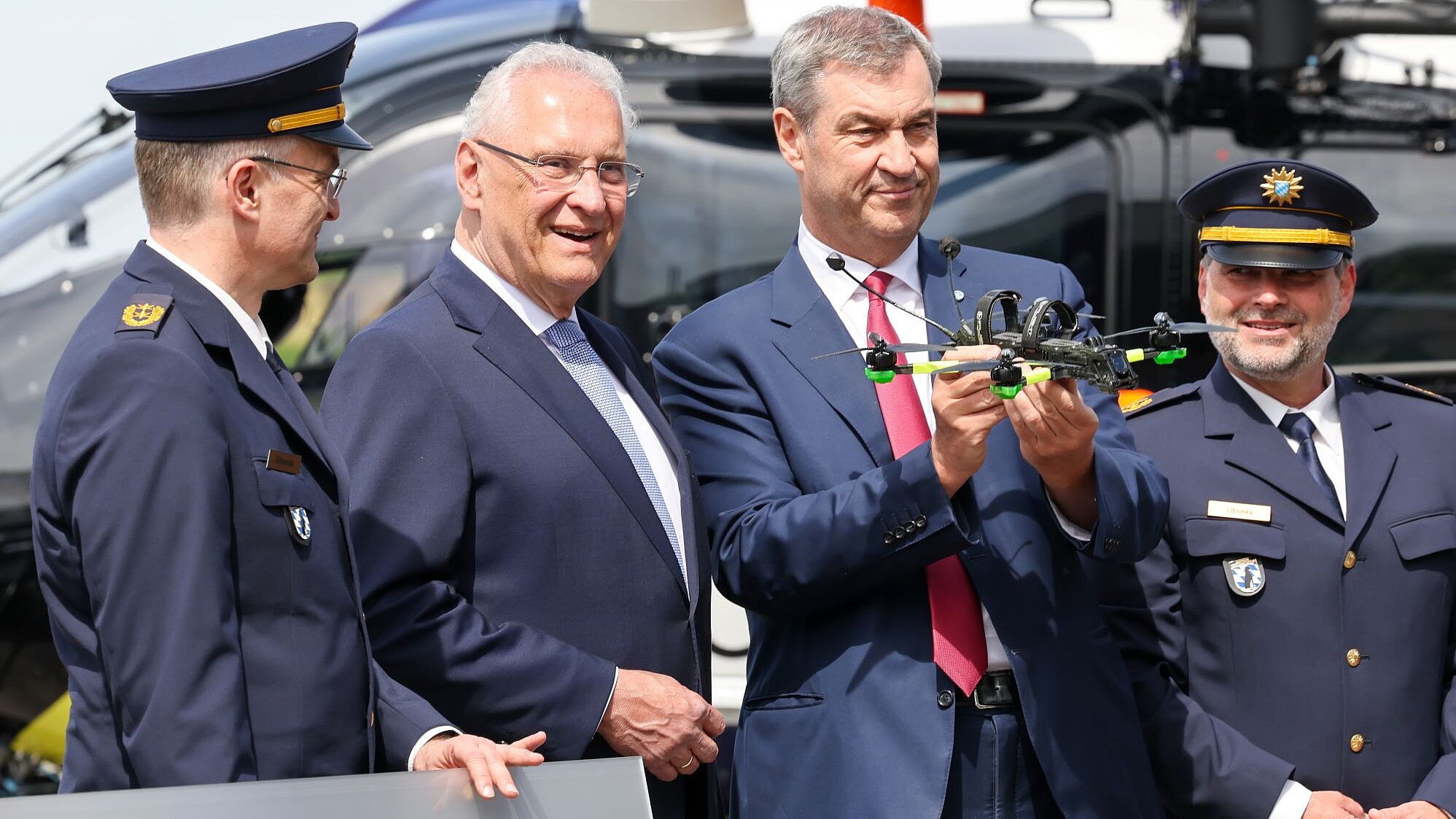 Innenminister Herrmann und Ministerpräsident Söder vor einem Hubschrauber, neben ihnen zwei Polizisten in Dienstuniform. Söder hält eine Mini-Drohne in der Hand.