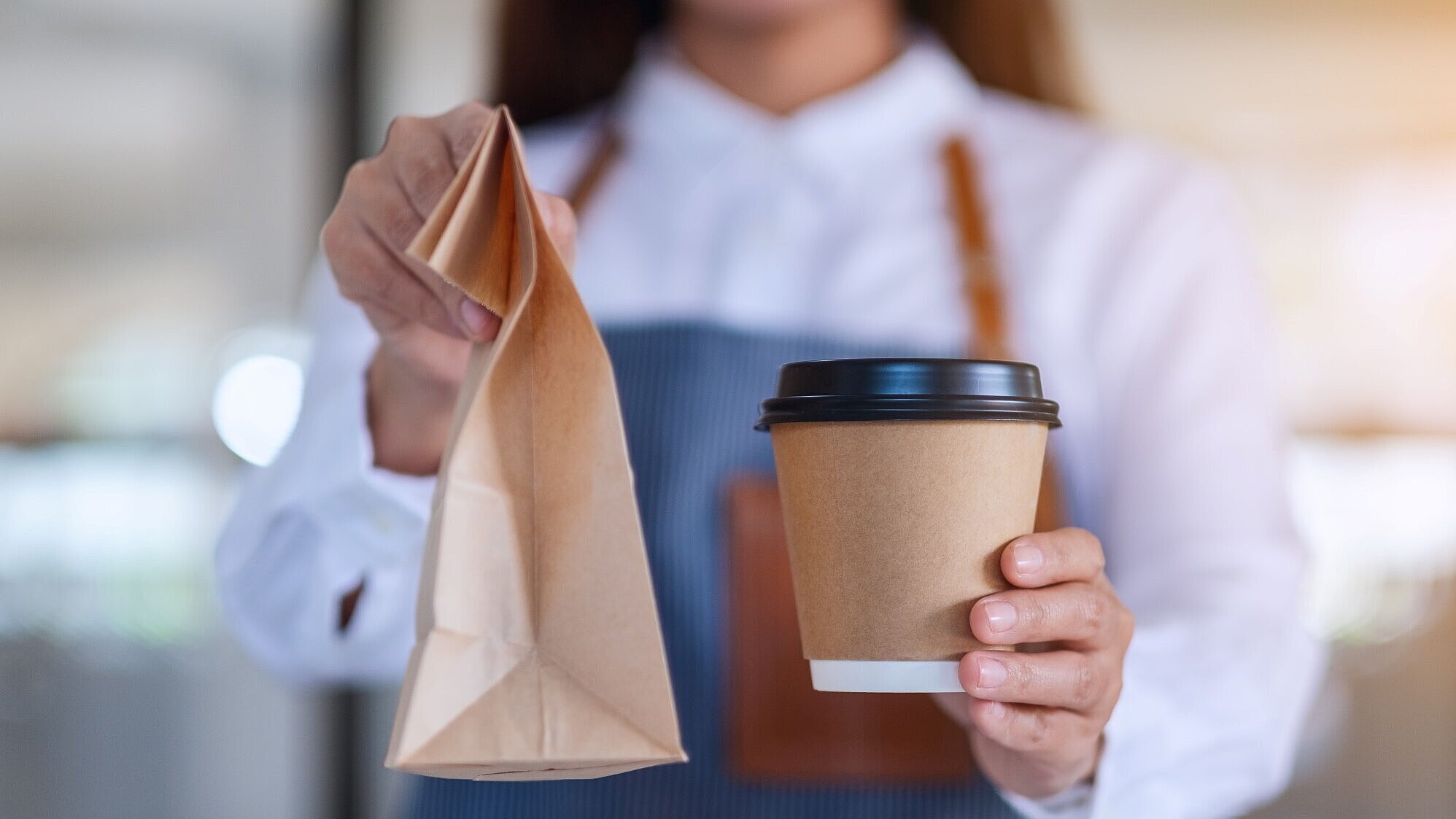 Frau mit Schürze hält einen Coffee-to-go-Becher und eine Papiertüte