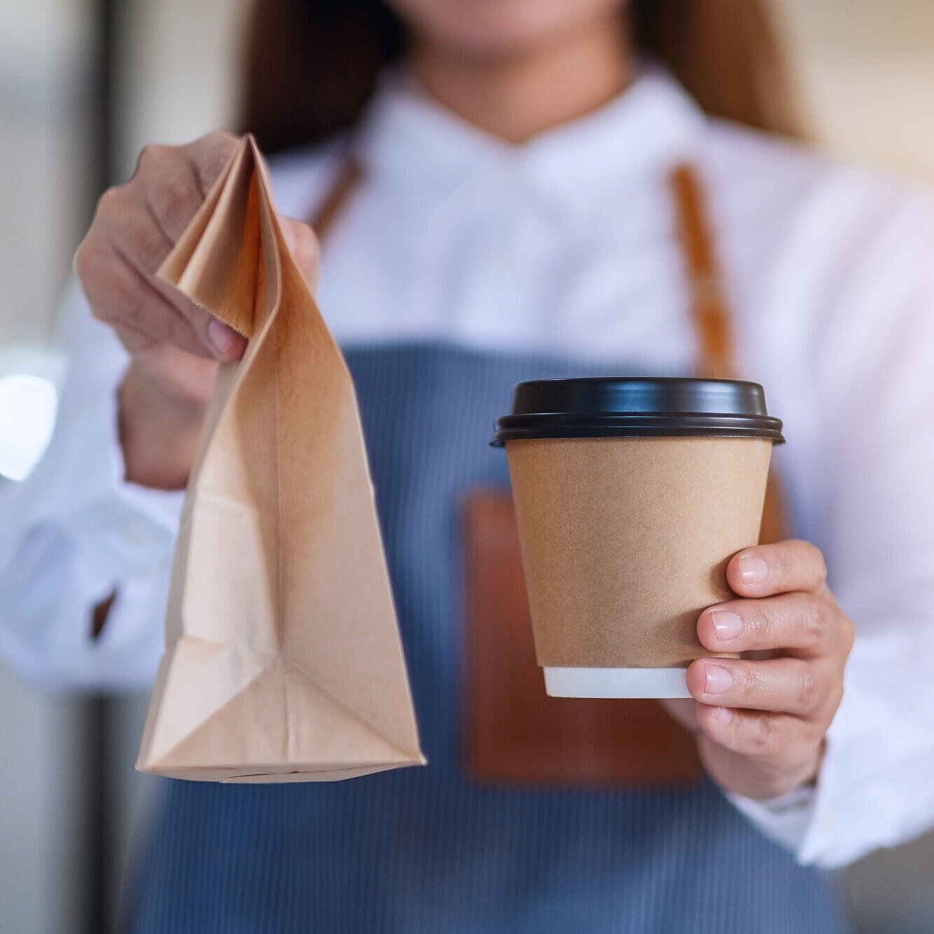 Frau mit Schürze hält einen Coffee-to-go-Becher und eine Papiertüte