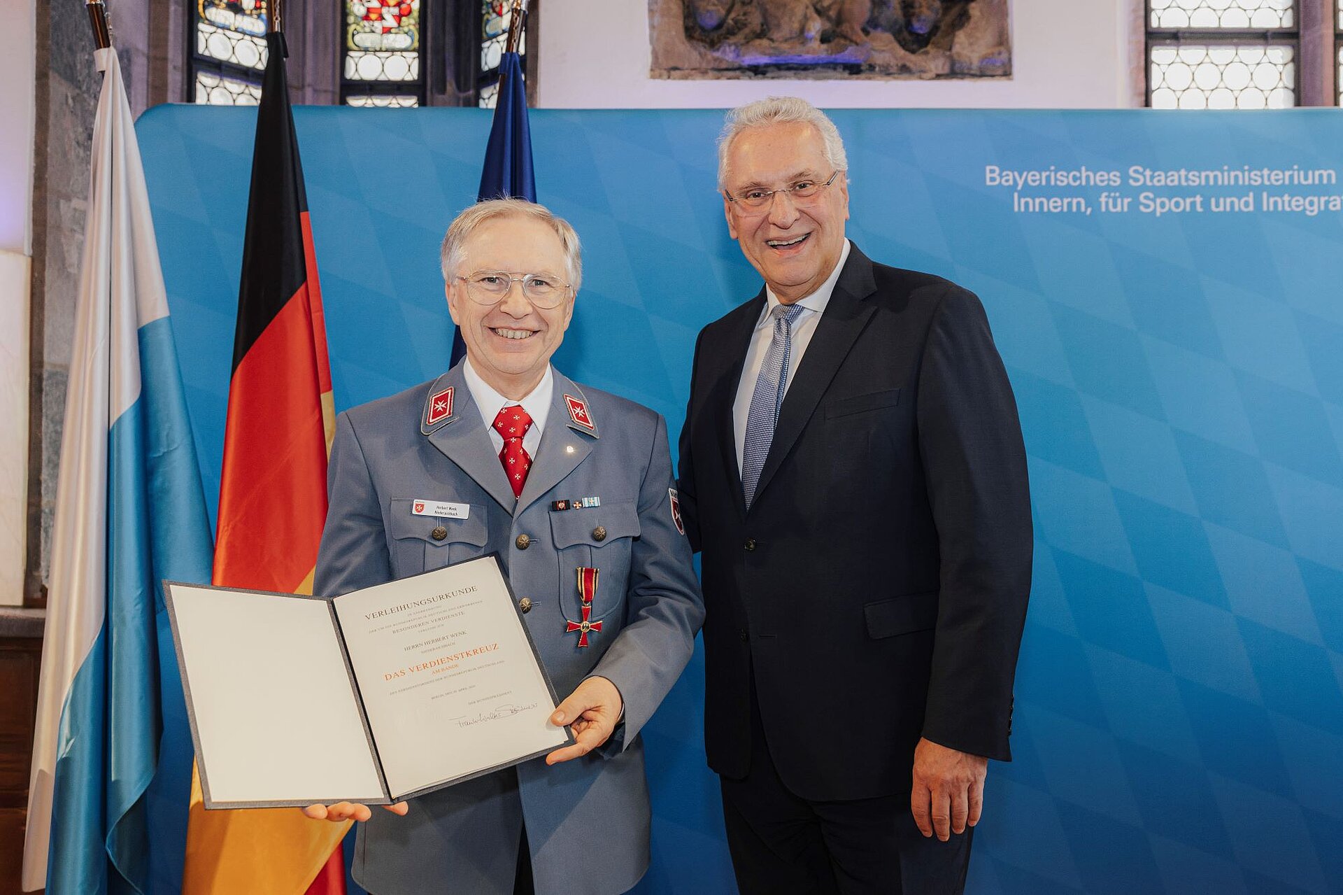 Innenminister Joachim Herrmann mit Herbert Wenk, der mit dem Verdienstkreuz am Bande ausgezeichnet worden ist. Hinter ihnen drei gehisste Fahnen sowie die Pressewand des Innenministeriums.