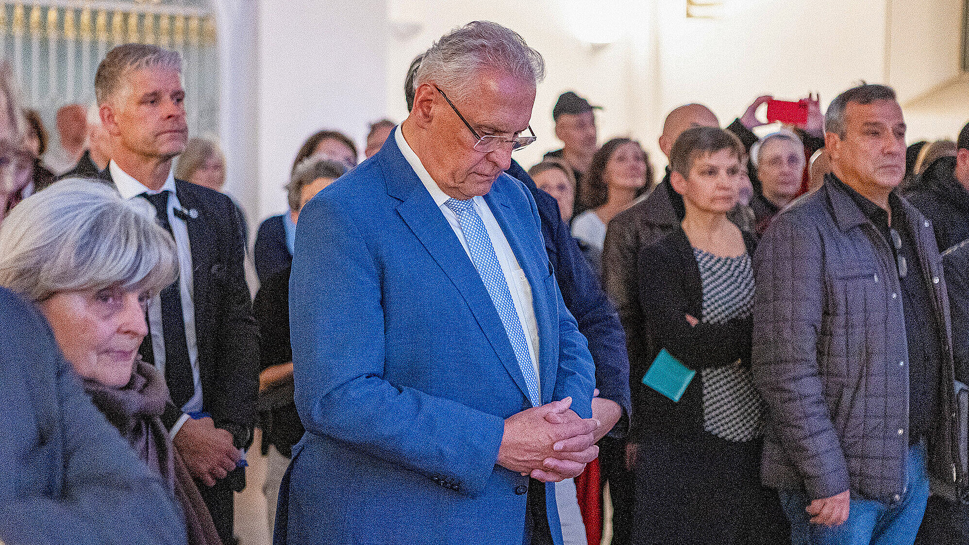 Das Bild zeigt Innenminister Joachim Herrmann und eine Gruppe von Menschen, die bei einer Veranstaltung in einem Raum versammelt sind und den Kopf zum Gebet gesenkt haben. Die Personen tragen formelle Kleidung, und es scheint sich um eine feierliche oder bedeutende Zusammenkunft zu handeln.
