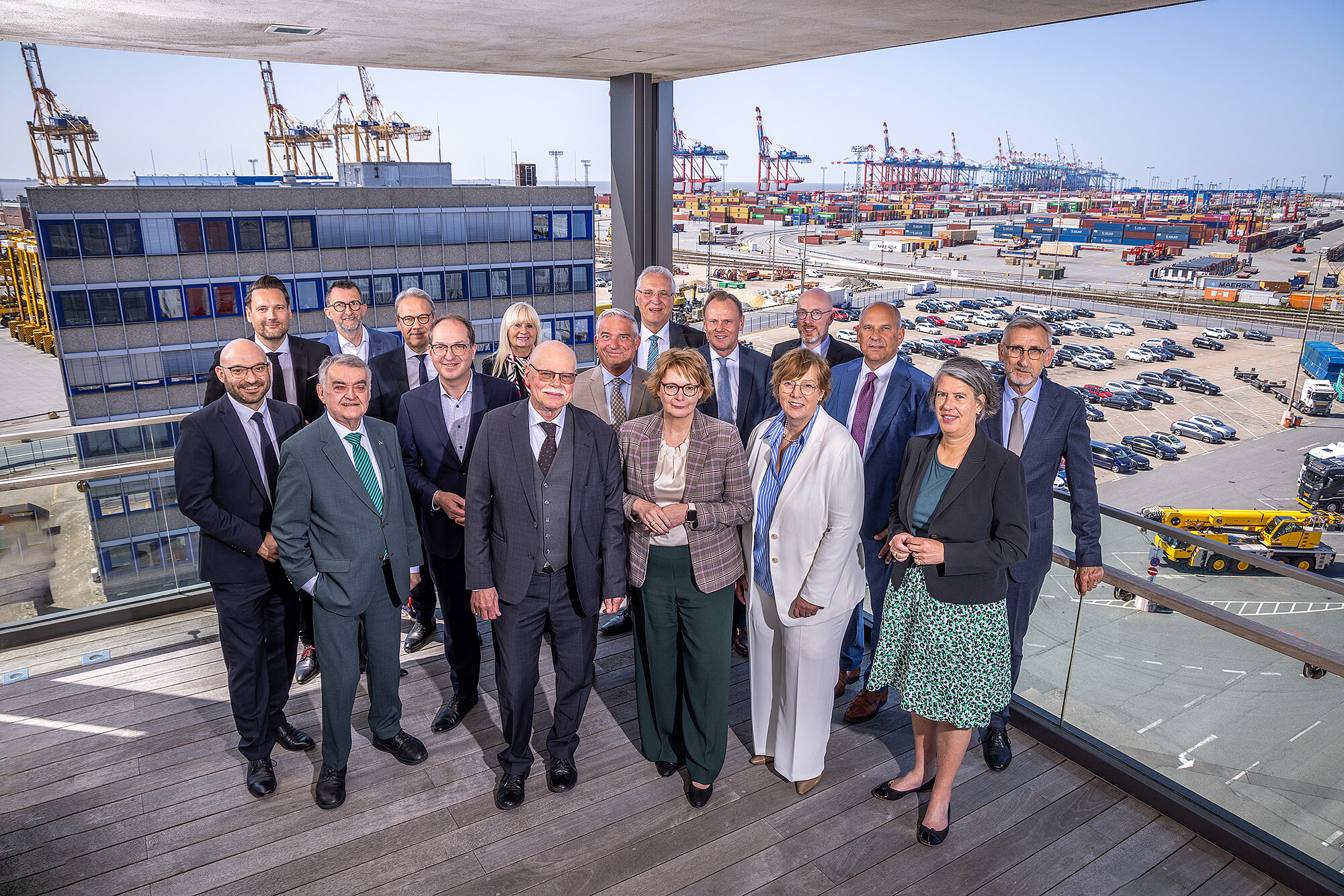 Gruppenfoto der Teilnehmer der IMK Personen in Businesskleidung auf einem Balkon mit Blick auf einen Containerhafen. Im Hintergrund sind zahlreiche Container, Kräne und geparkte Autos zu sehen. Die Personen stehen eng beieinander und lächeln in die Kamera. 
