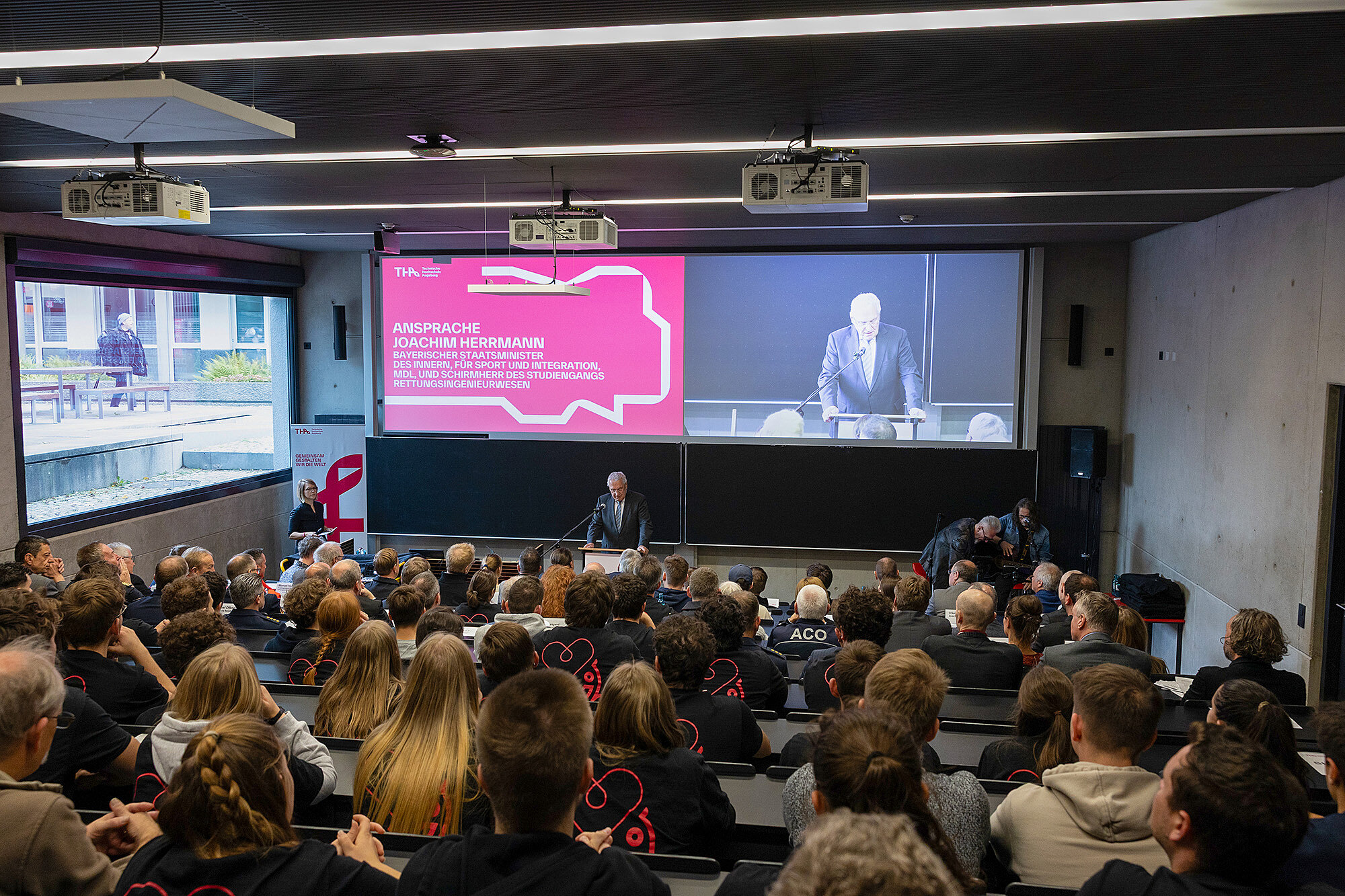 Ein Bild von einer Veranstaltung im Hörsaal, bei der Innenminister Joachim Herrmann am Rednerpult spricht. Auf der großen Leinwand im Hintergrund ist eine Präsentation mit dem Titel "Ansprache Joachim Herrmann" zu sehen. Der Saal ist voll mit Zuhörern, die aufmerksam dem Vortrag folgen.