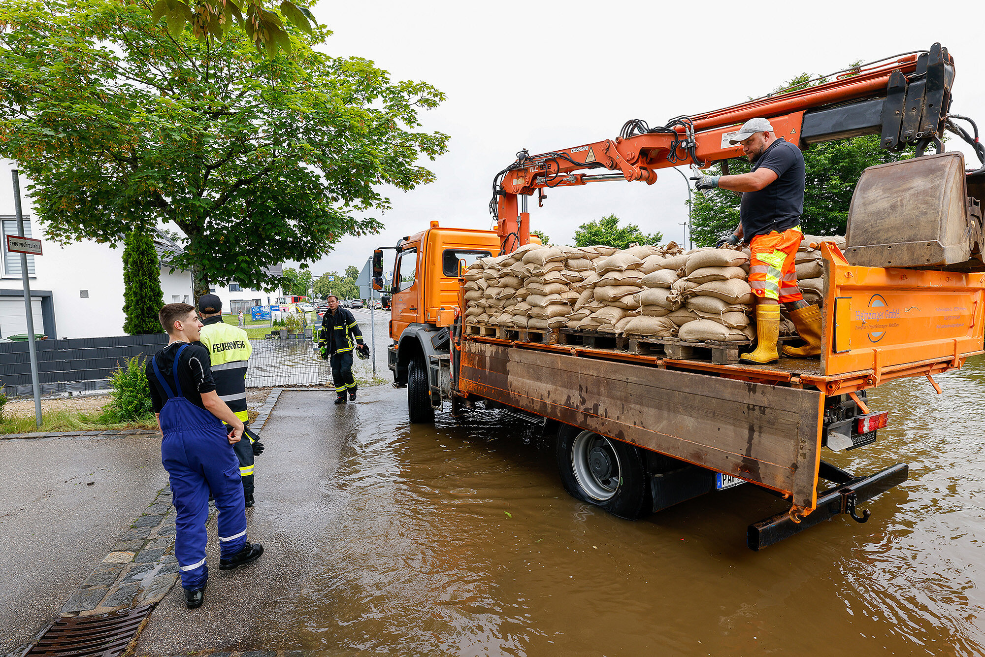 Das Bild zeigt einen Einsatz bei starkem Regen, bei dem Feuerwehrkräfte und Helfer mit einem orangefarbenen Einsatzfahrzeug auf einer überschwemmten Straße arbeiten. Ein Mann steht auf dem Fahrzeug, das mit Sandsäcken beladen ist, und überprüft die Ausrüstung. Zwei weitere Helfer in Feuerwehruniformen stehen am Straßenrand, während Wasser die Straße überschwemmt. Im Hintergrund sind Bäume, Gebäude und Einsatzfahrzeuge zu sehen.