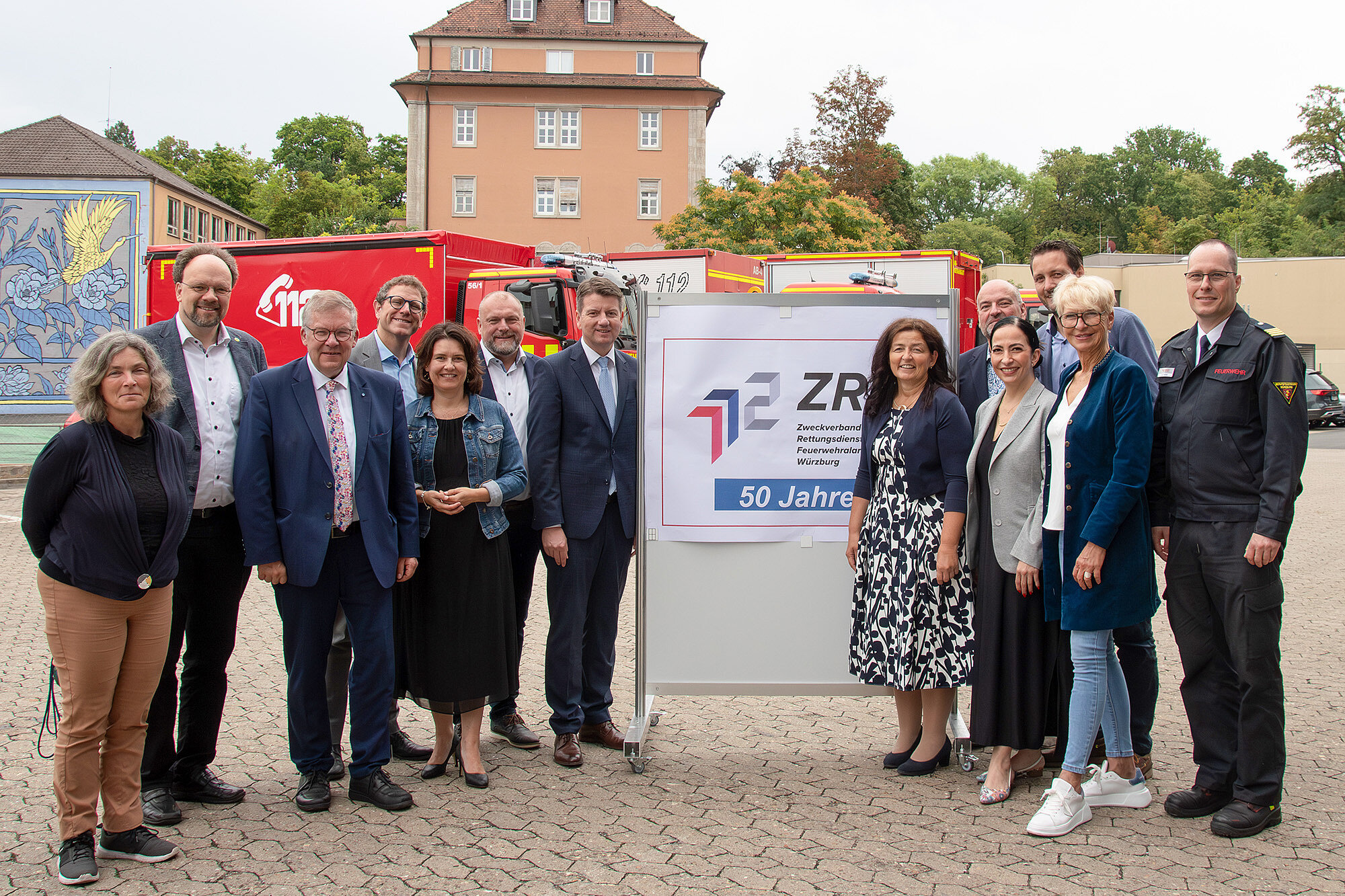 Gruppenbild von links nach rechts Ltd. Branddirektor Harald Rehmann, Landrat Thomas Eberth, Staatssekretär Sandro Kirchner, stv. Landrätin und Vorsitzende des ZRF Würzburg Christine Haupt-Kreutzer, Geschäftsführer des ZRF Würzburg Paul Justice vor einem roten Feuerwehrauto im Hintergrund ein Haus und ein Plakat auf dem ZRF steht.