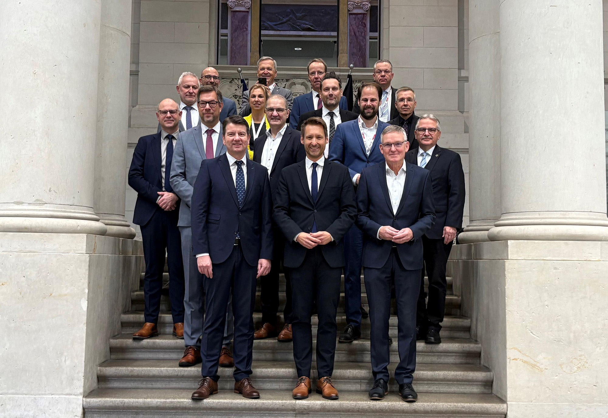 Gruppenfoto von 17 Personen (1 v. li. erste Reihe Staatssekretär Sandro Kirchner), die in mehreren Reihen auf einer Treppe vor einem Gebäude mit großen Säulen stehen. Die Personen tragen überwiegend dunkle Anzüge und Hemden, einige haben Krawatten an. Im Hintergrund sind zwei Fahnen zu sehen.